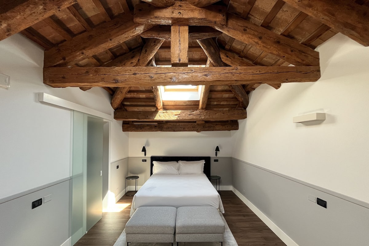 Ein schmaler Flur, der in ein Schlafzimmer mit einem großen Bett, einer Bank und einem Oberlicht führt.