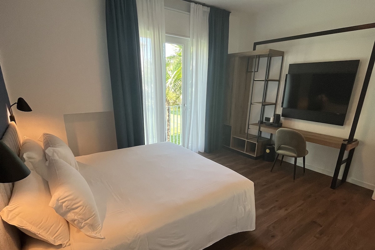 Ein modernes Hotelzimmer mit einem großen Bett, einem Schreibtisch, einem Sessel, einem Fernseher und einem Balkon mit Vorhängen.
