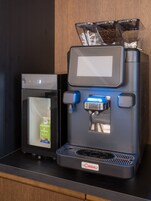 Una cafetera con espumador de leche y dispensador de leche, un molinillo de café y un recipiente de vidrio con granos de café.
