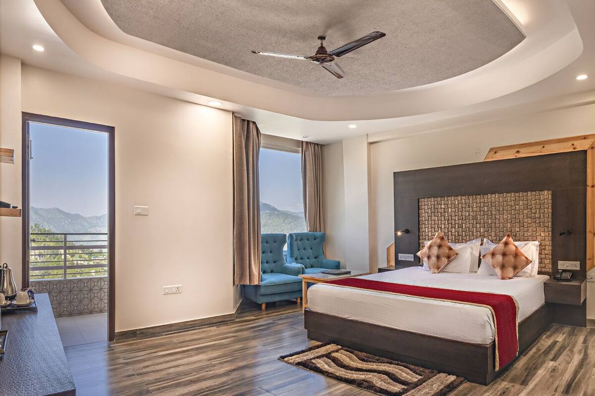 Une chambre d’hôtel moderne avec un grand lit, un coin salon doté d’un fauteuil bleu, un ventilateur de plafond et une vue sur les montagnes depuis le balcon.