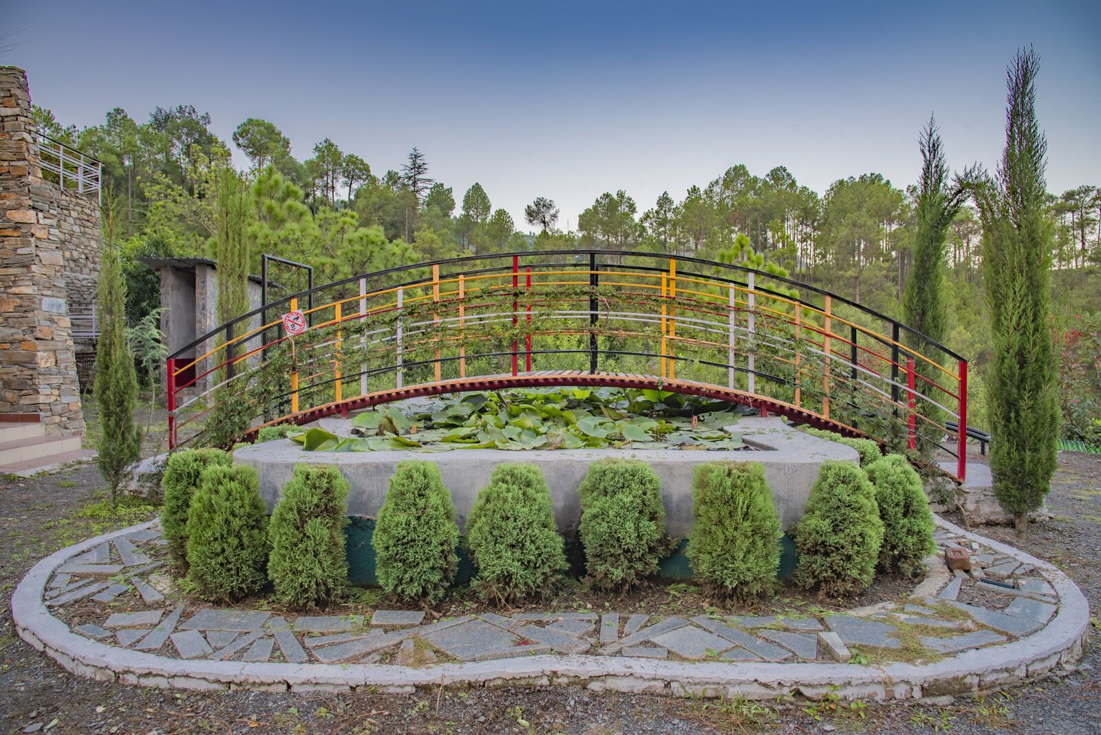 A stone bridge with a metal railing, surrounded by a circular garden with trimmed bushes and a pond with lily pads.