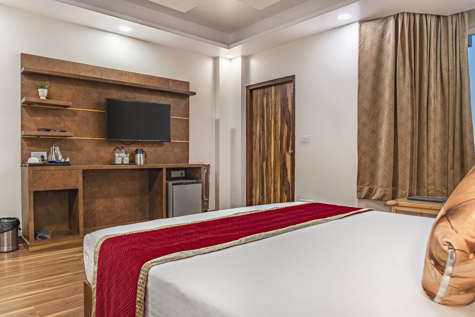 A hotel room with a bed, a TV mounted on a wooden wall, a desk with a computer, a small fridge, and a wardrobe.