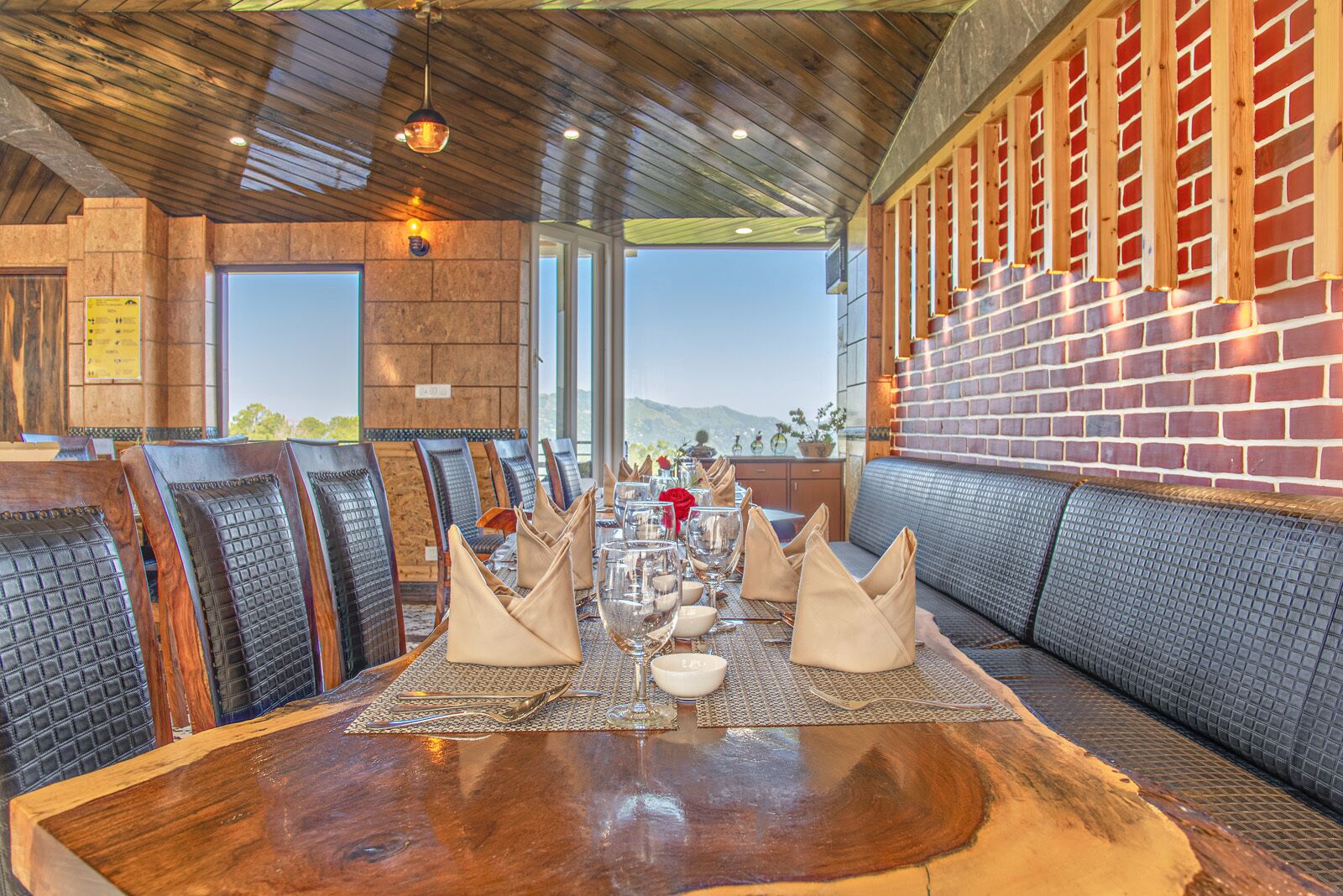 A restaurant with a wooden table set for dining, cushioned seating, and a brick wall with windows offering a view.