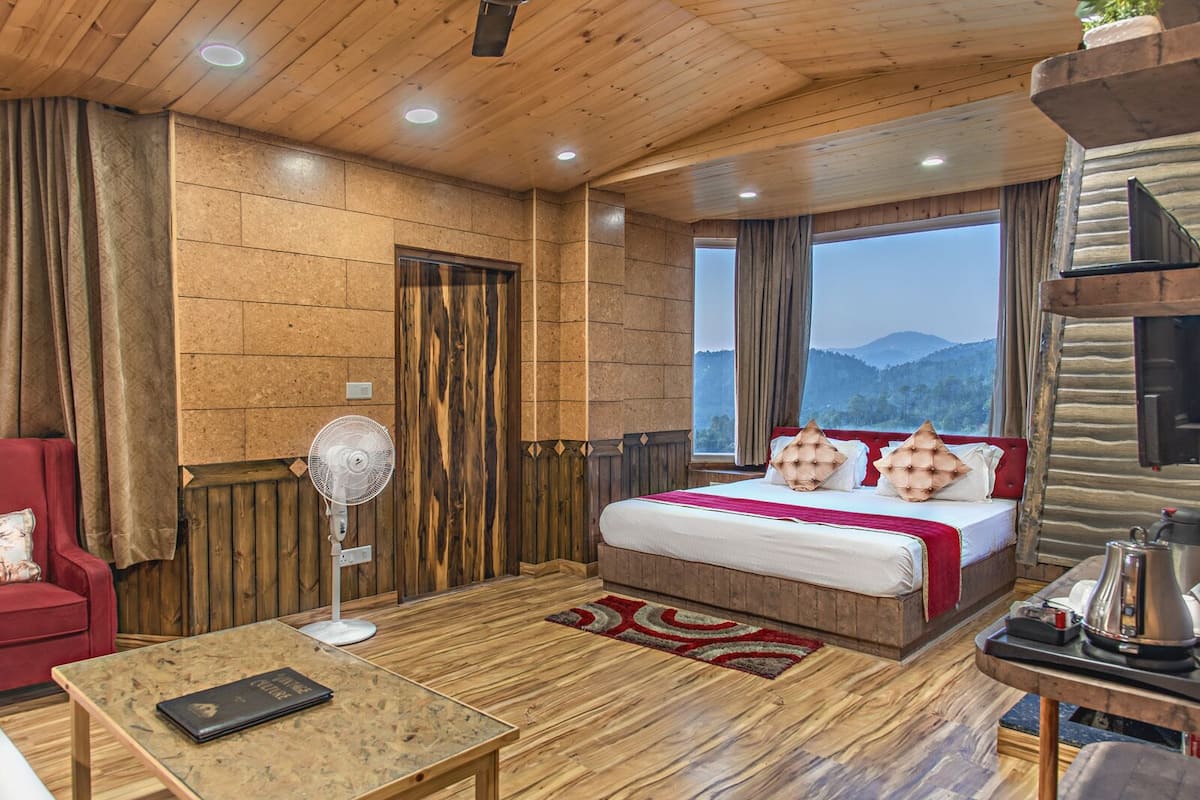 Une pièce chaleureuse avec un plancher en bois, un lit avec une literie rouge, un fauteuil rouge, une table avec un livre et une grande fenêtre donnant sur les montagnes.