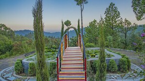 A wooden bridge with colorful stripes leading to a circular garden area with a pond and surrounding greenery. - Kanishka Retreat Resort Chail (Kandaghat)