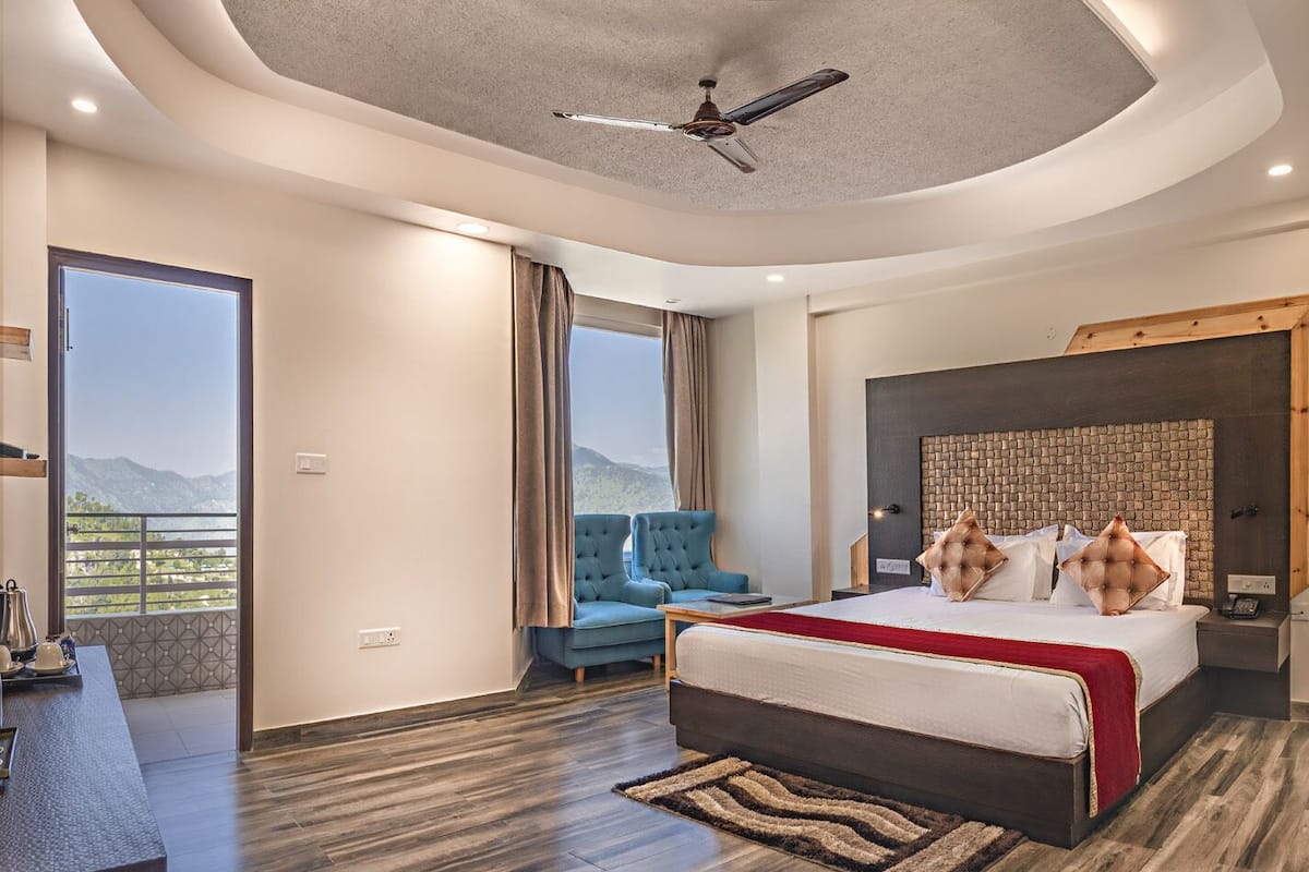 Une chambre d’hôtel moderne avec un grand lit, un coin salon avec un fauteuil bleu, un ventilateur de plafond et une vue sur les montagnes.