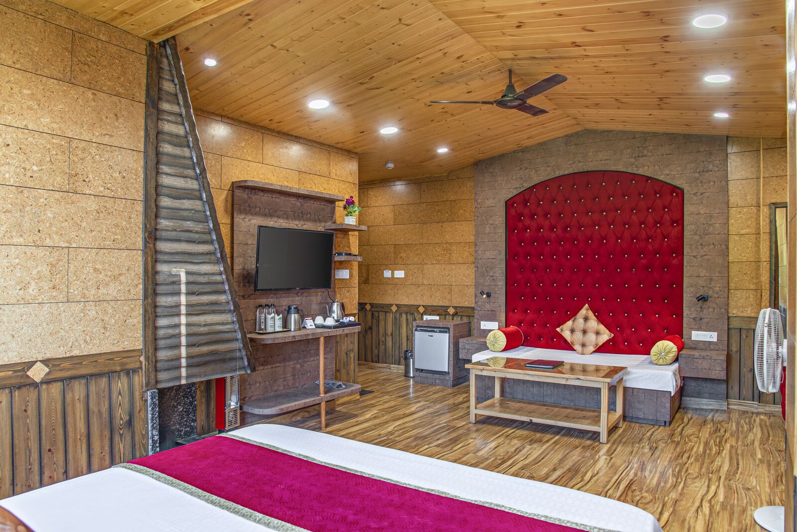 A cozy room with a wooden ceiling, a red upholstered headboard, a television mounted on the wall, a small table with a red cushion, and a bed with a pink blanket.