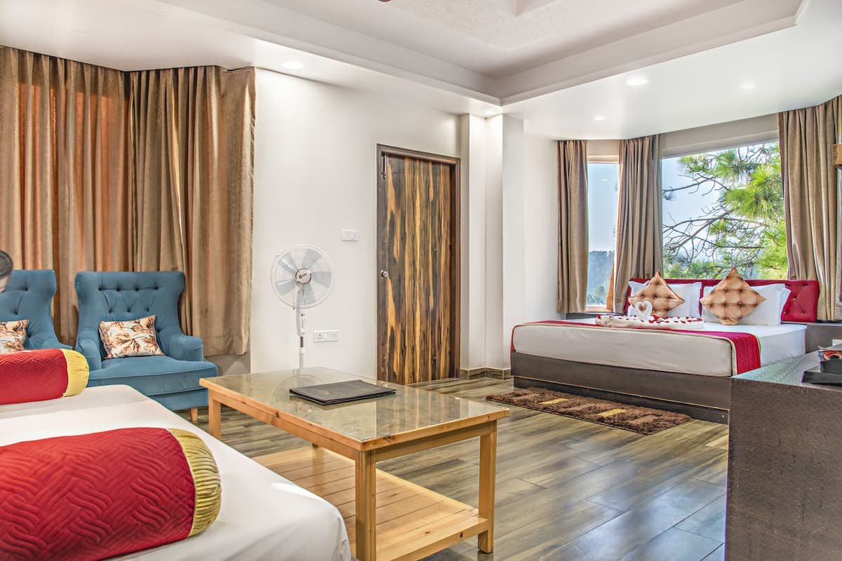 Une chambre d’hôtel avec un lit, deux fauteuils bleus, une table basse en bois, un ventilateur et une grande fenêtre avec des rideaux.