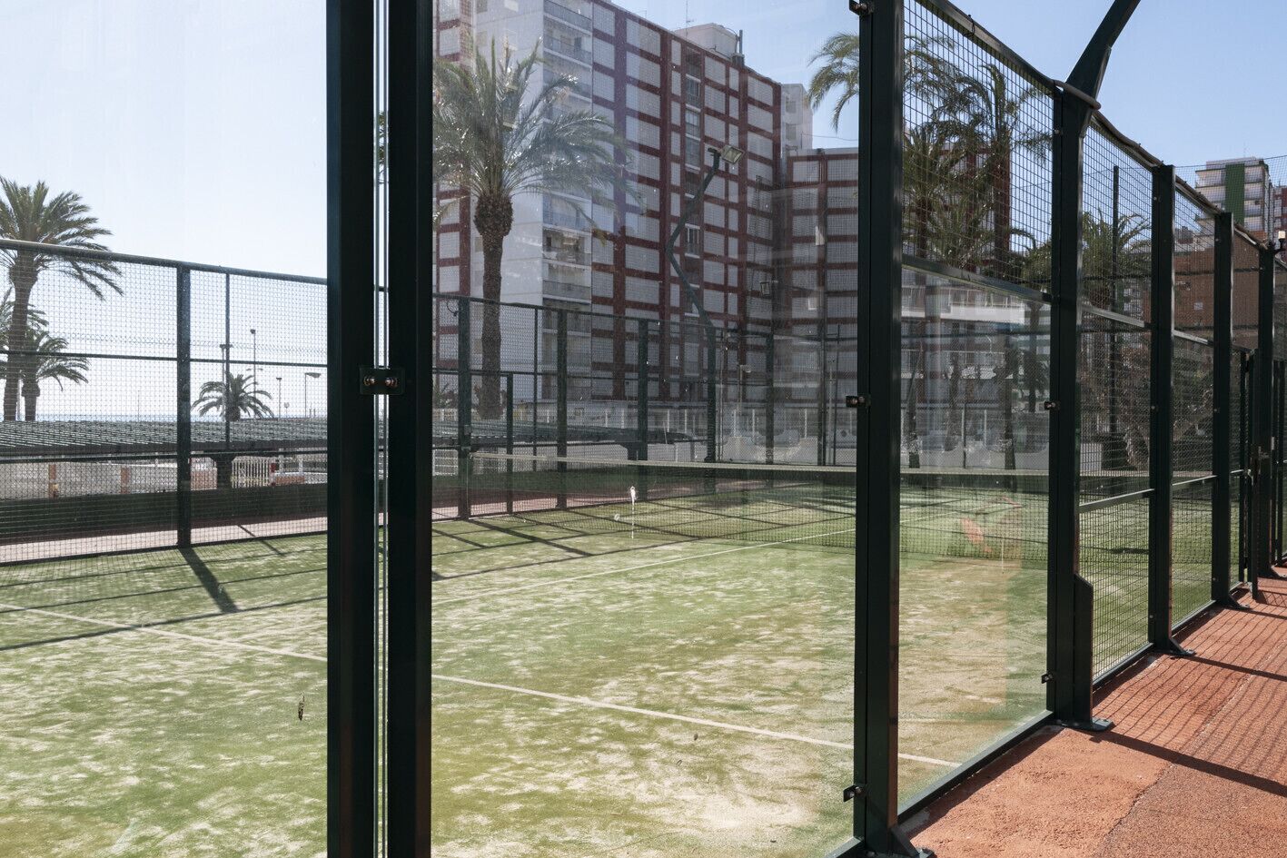 Un court de tennis entouré d’une clôture métallique, avec en arrière-plan la vue d’un immeuble de grande hauteur.
