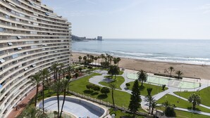 Ein Hochhaus-Hotel mit mehreren Balkonen mit Blick auf einen Strand und einen Poolbereich mit Palmen und einem Tennisplatz.