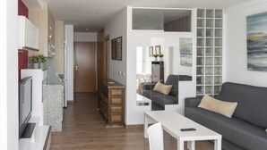 Ein modernes Wohnzimmer mit grauem Sofa, weißem Couchtisch und Fernseher. Es gibt eine Küchenzeile mit Holzschränken und einem Kühlschrank.