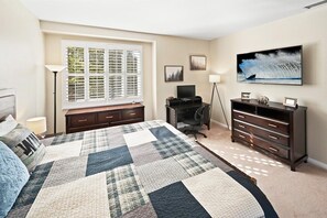 A bedroom with a bed, a desk, a dresser, a television, and a window with blinds.