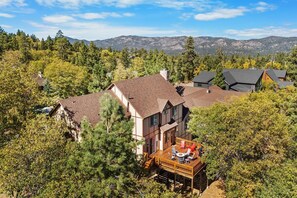 A two-story house with a deck, surrounded by trees and mountains in the background. - High Timber Family Retreat: Luxury Family Fun in High Timber Ranch! Spacious Fenced Yard! Game Room! (Big Bear City)