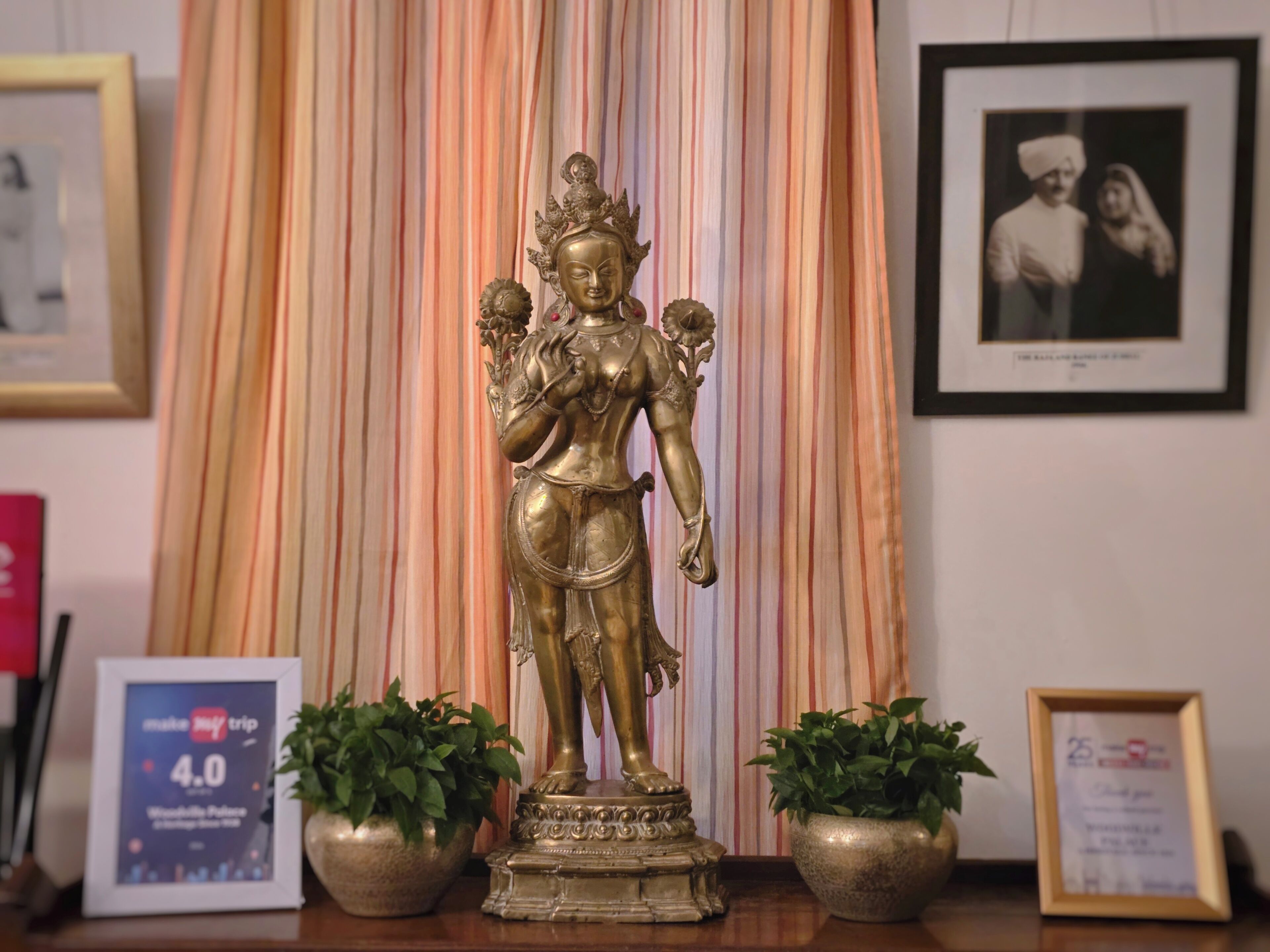 A statue of a deity with two potted plants on either side, framed pictures on the wall, and a framed certificate on the table.