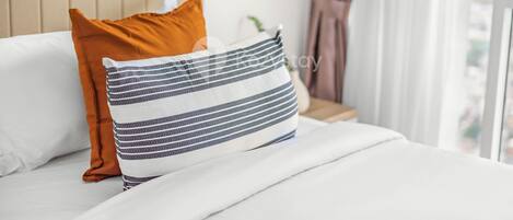 A neatly made bed with white and striped pillows, a beige headboard, and a view of a cityscape through a window with curtains.