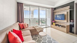 A modern living room with a large flat-screen TV, a sofa with red cushions, a small round table, and a sliding glass door leading to a balcony. - SOLEIL by Kozystay (Jakarta)