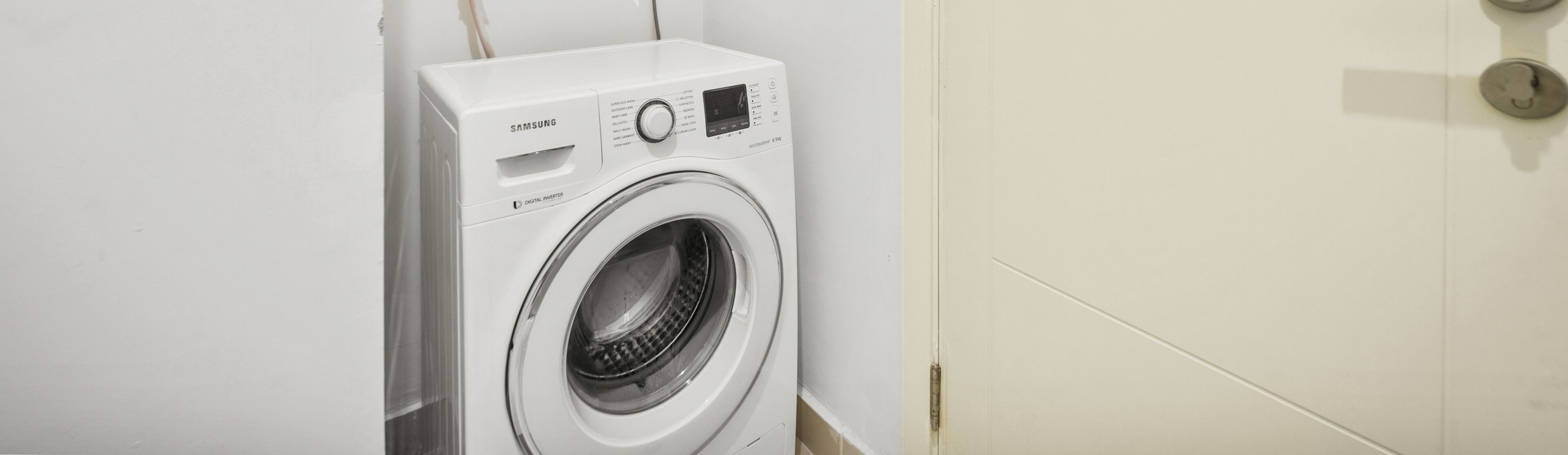 Un lave-linge Samsung blanc, doté d’un écran numérique et d’un panneau de commande, se trouve dans une pièce aux murs unis et avec une porte.