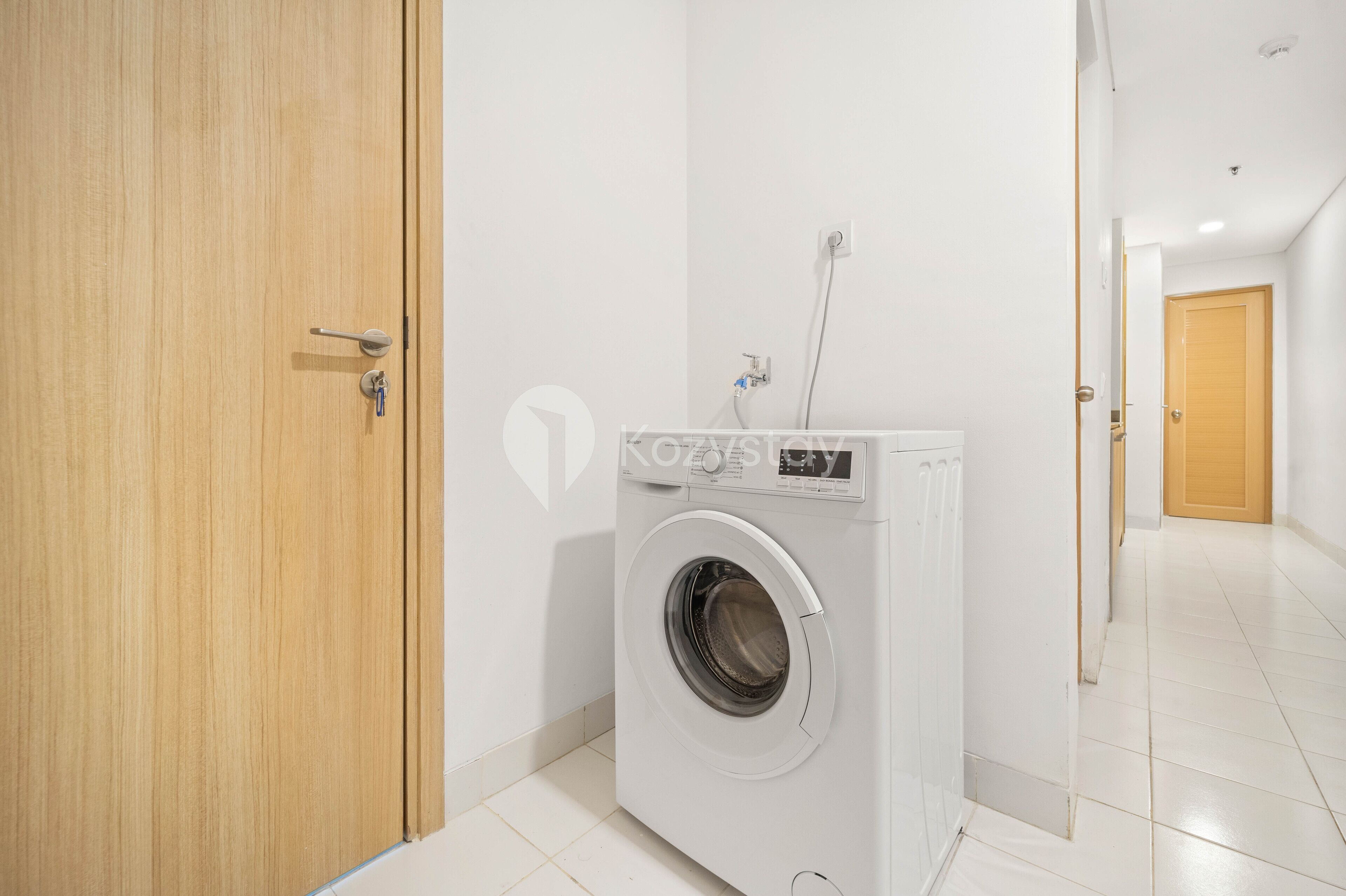 A laundry room with a front-loading washing machine, a wooden door, and a hallway leading to another room.