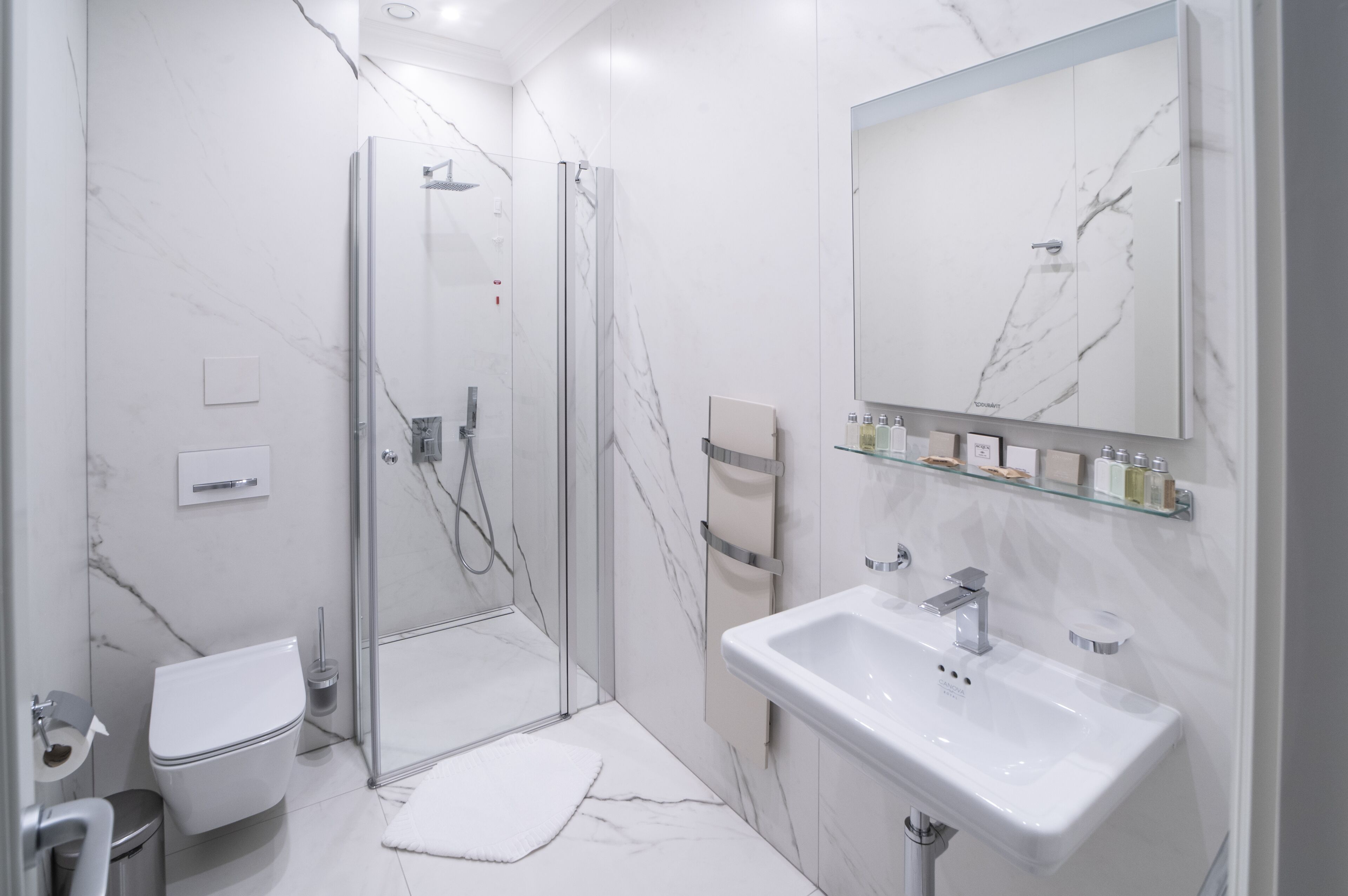 A modern bathroom with a glass shower enclosure, a white toilet, a sink with a mirror above it, and a marble-patterned wall.