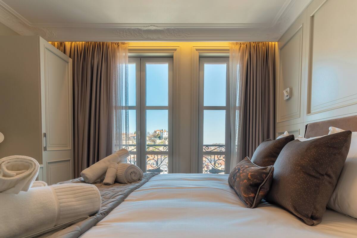 Quarto de hotel com cama, almofadas, toalha enrolada e vista para a cidade através de grandes janelas.