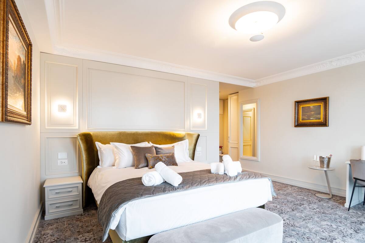 Quarto de hotel com uma cama grande, mesas de cabeceira, uma mesinha e uma cadeira.