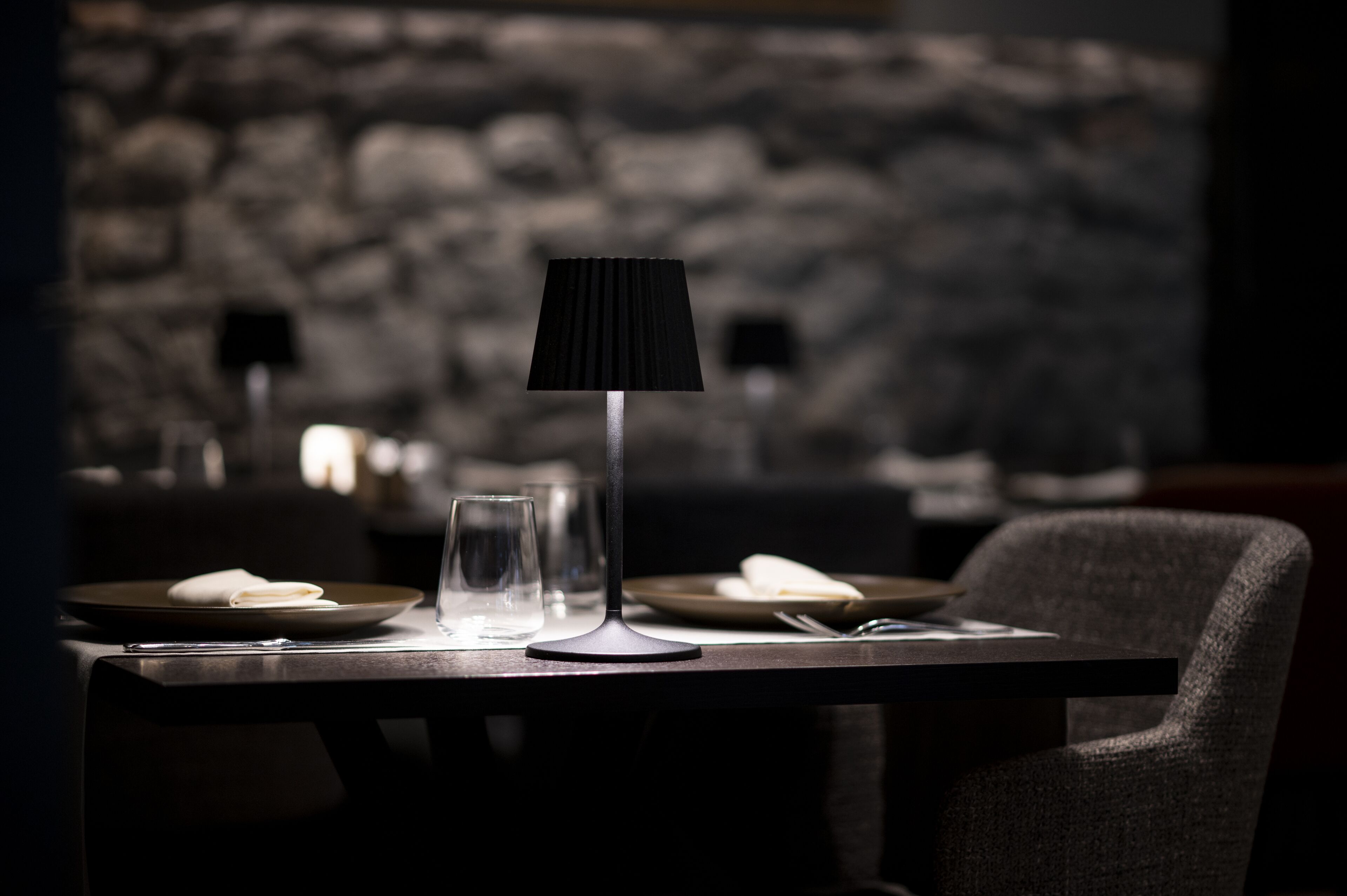 Un comedor tenuemente iluminado con una mesa dispuesta para dos, que incluye una lámpara moderna, platos, vasos y una servilleta doblada.
