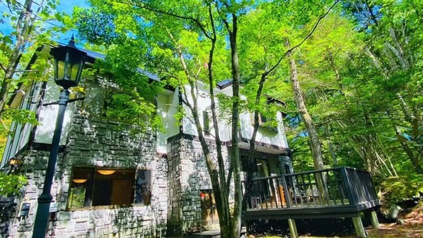 A rustic stone building with a wooden deck, surrounded by lush greenery and trees. - IPPUKAKU (Tsumagoi)