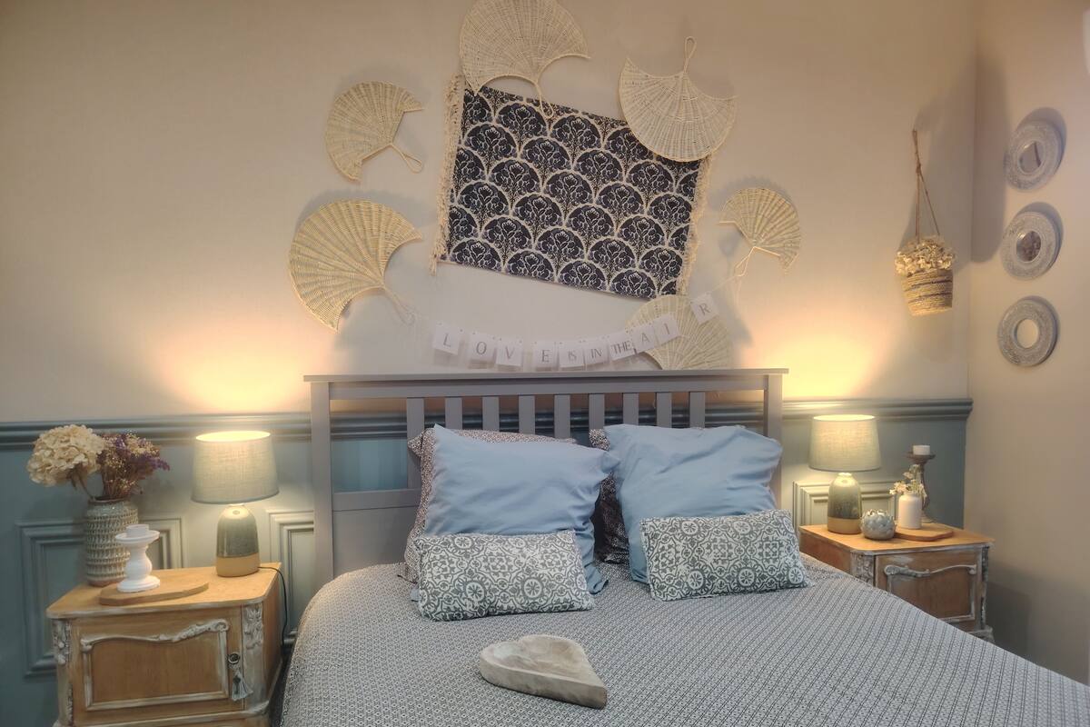 Ein Schlafzimmer mit einem Bett, zwei Nachttischen, einer Wandlampe, Wanddekoration und einer Vase auf dem Nachttisch.