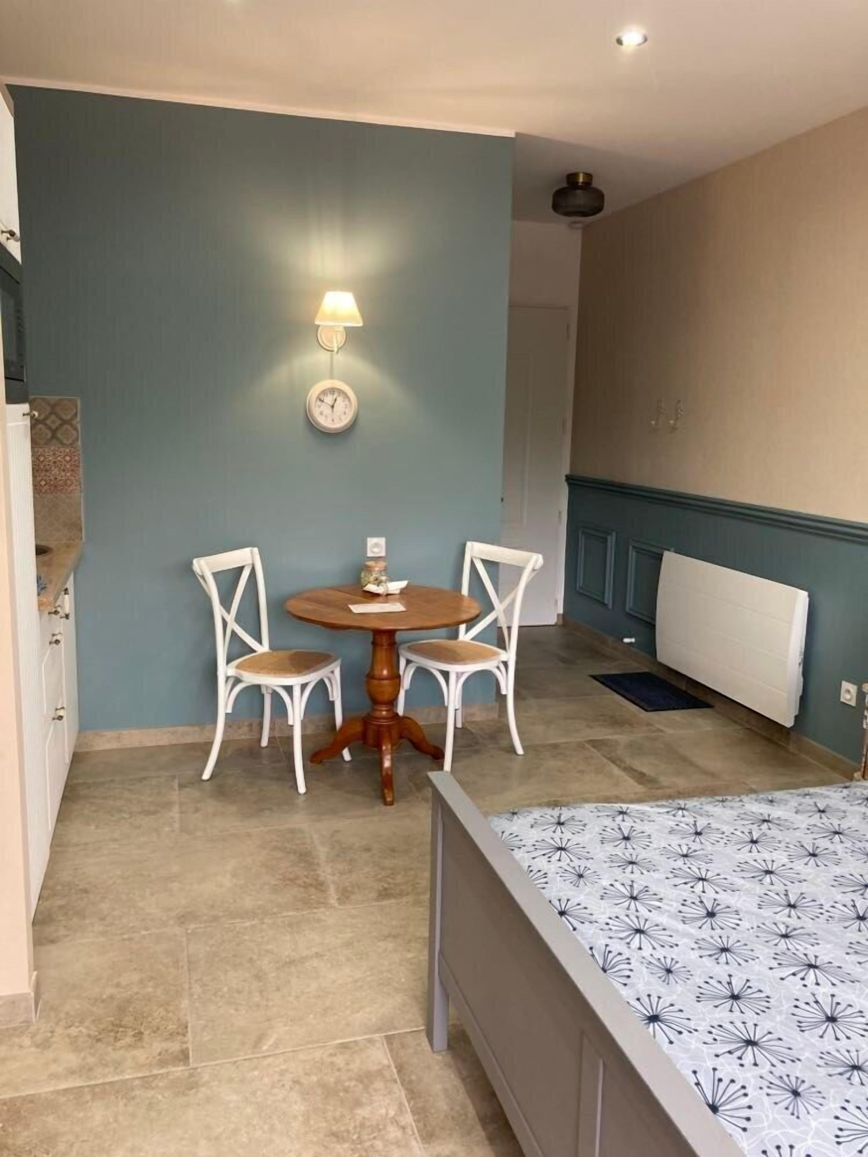 A compact room with a bed, a small round table with chairs, a wall clock, and a radiator.