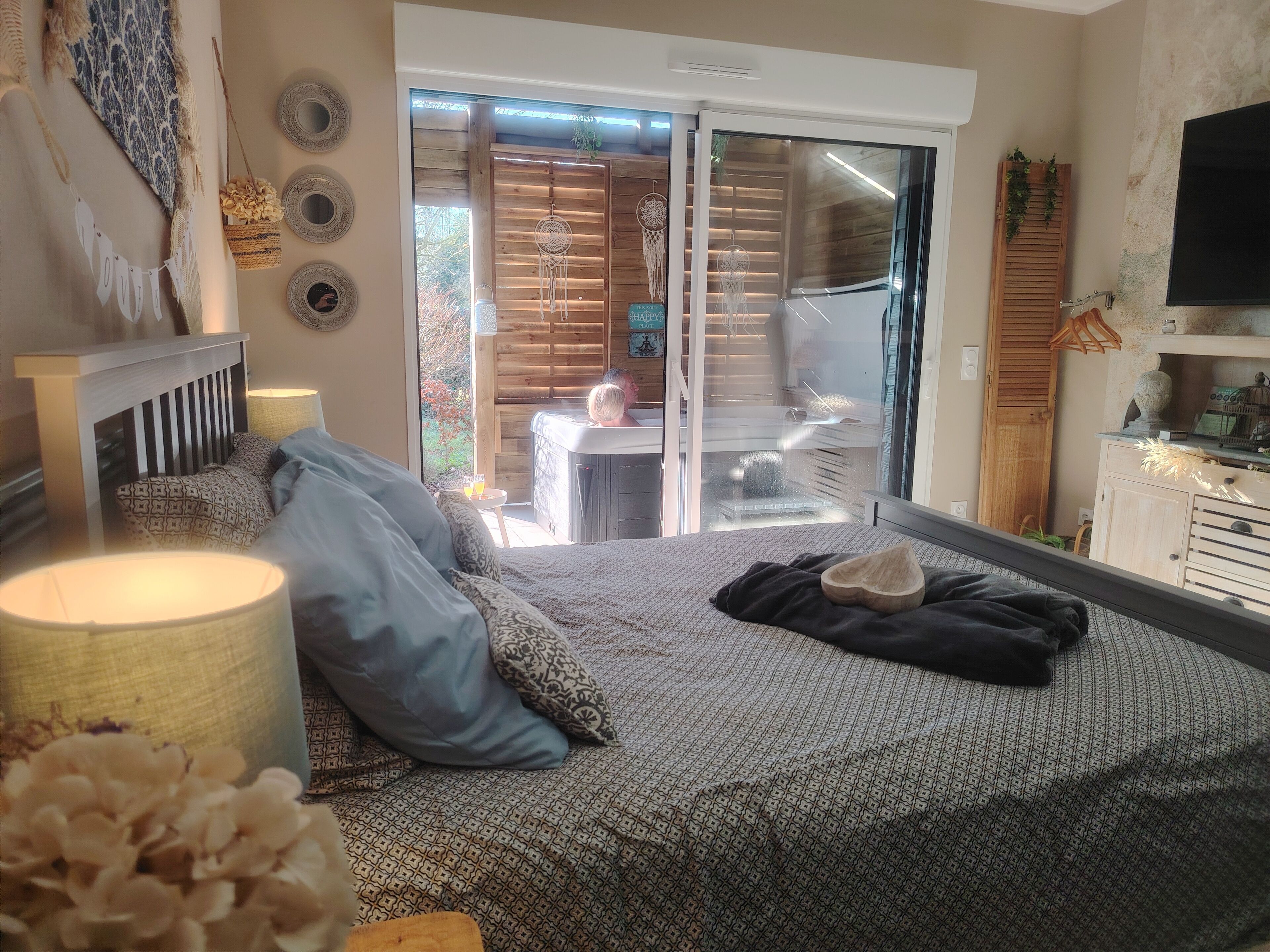 Een slaapkamer met een bed, kussens, een bedlampje, een houten hoofdbord, een schuifdeur van glas die leidt naar een balkon, en een aan de muur bevestigde televisie.