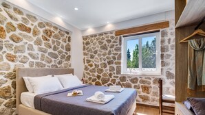 A bedroom with a stone wall, a bed with a quilt, a window with a view, and a wooden chair. - Apartment "Superior Apartment A" close to the Beach with Shared Pool & Wi-Fi (?e???da)