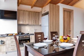 A modern kitchen with wooden cabinets, a refrigerator, and a dining table set for two with wine glasses and fruit.