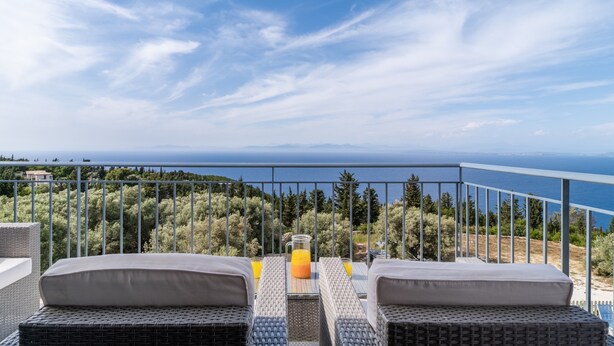 Uma varanda com móveis de vime, uma mesa de vidro e vista para o mar e as árvores.