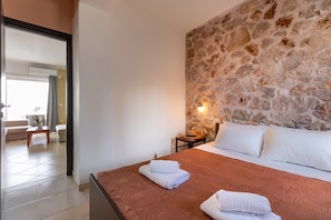 A hotel room with a stone wall, a bed with white linens, a nightstand with a lamp, and a view of the living area.