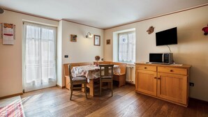 A room with a wooden dining table, chairs, a television on a cabinet, and a window with curtains. - Holiday Apartment 'Le Coccinelle' with Mountain View, Terrace & Wi-Fi (Varena)