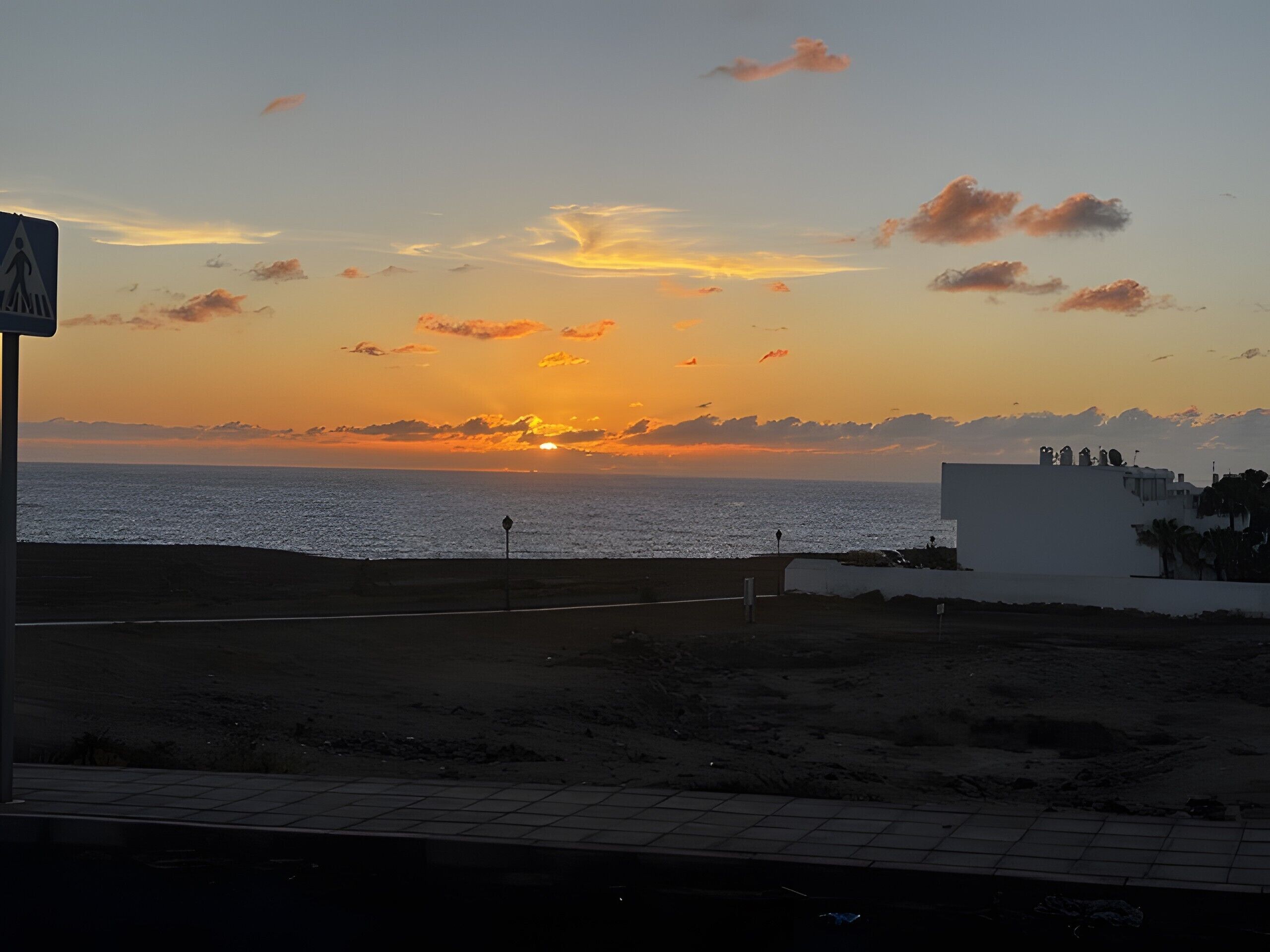 A sunset over the sea with a pedestrian crossing sign, a paved walkway, and a building with a flat roof.