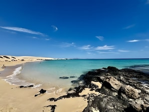Una playa con agua turquesa cristalina, una orilla de arena y afloramientos rocosos.