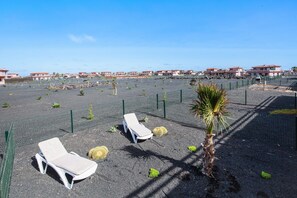 Une plage de sable avec des chaises longues et un palmier, entourée d’une clôture et bordée de bâtiments en arrière-plan.