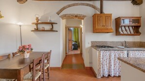 Une cuisine rustique avec une table en bois, des chaises, un luminaire suspendu et une arche en briques qui mène à une autre pièce.