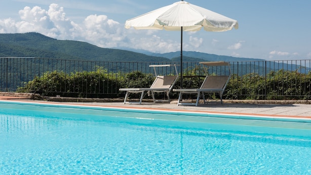 Une piscine entourée d’une clôture et de verdure, avec des chaises longues et un parasol, et des montagnes en arrière-plan.