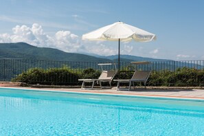 Une piscine entourée d’une clôture et de verdure, avec des chaises longues et un parasol, et des montagnes en arrière-plan.