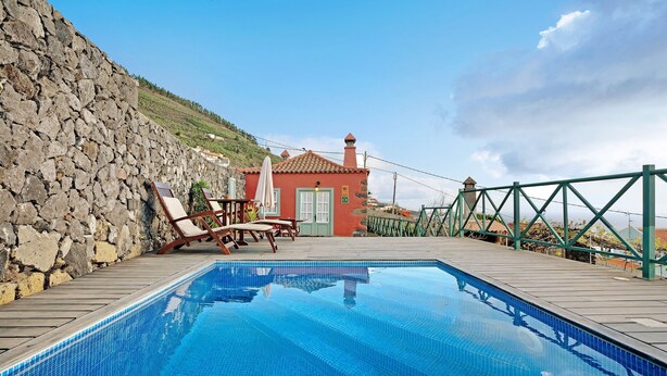Une piscine bordée d’un mur en pierre, avec des chaises longues et un petit bâtiment doté d’un toit en tuiles.