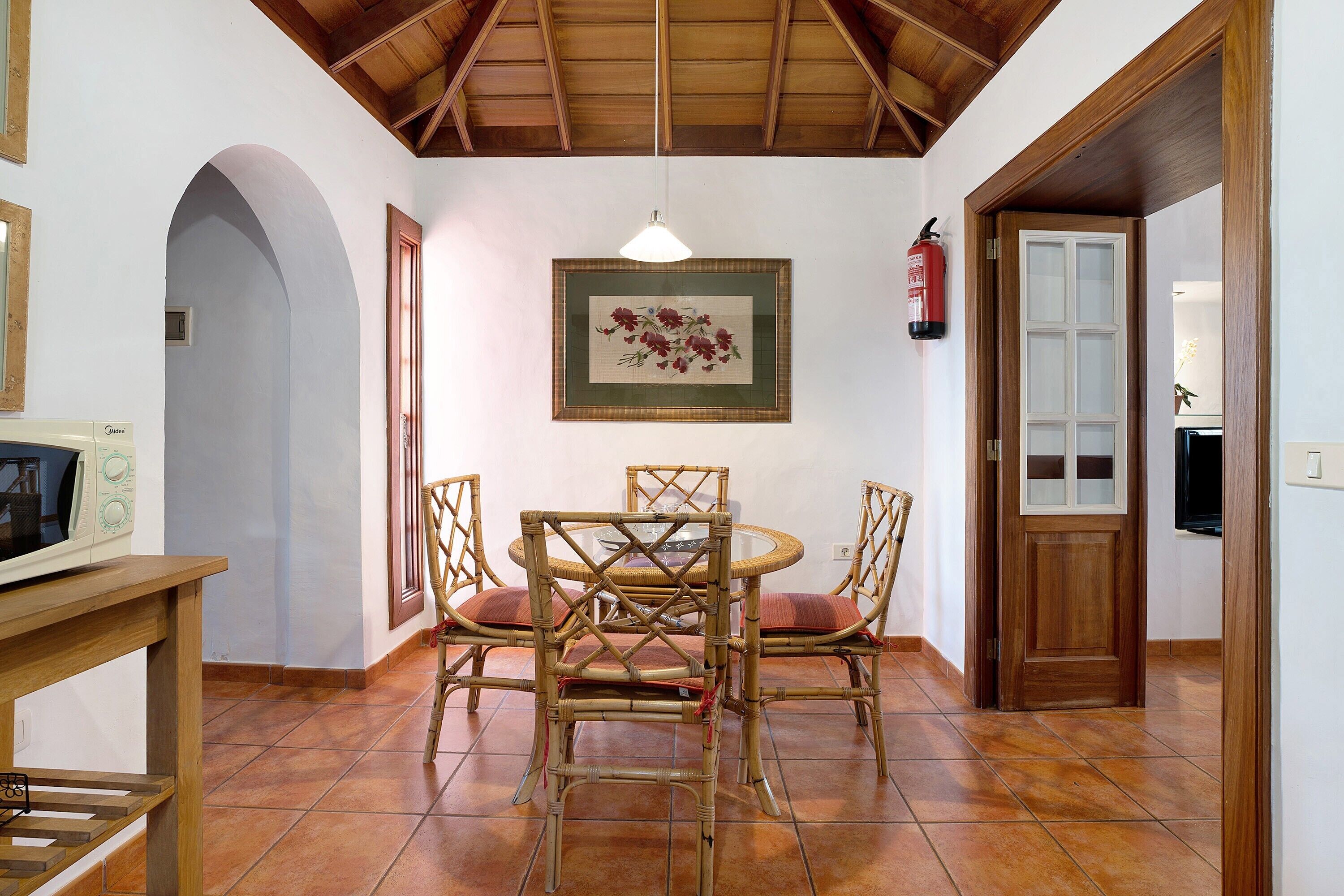 A dining area with a round table and four chairs, a framed floral artwork on the wall, a fire extinguisher, and a wooden door.