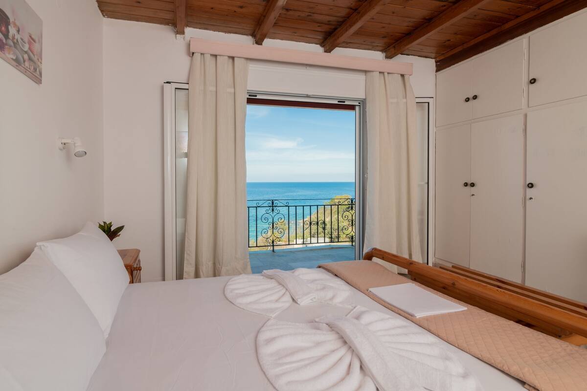 A bedroom with a bed, a wardrobe, a balcony with a view of the sea, and a painting on the wall.