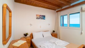 A bedroom with a wooden ceiling, a bed with white linens, a bedside table with a plant, and a window with a view of the sea. - Agnadi Sea View Apartments (Zakynthos)