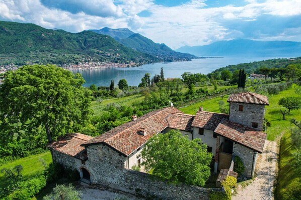 A historic stone building with a tiled roof, surrounded by lush greenery and a lake.