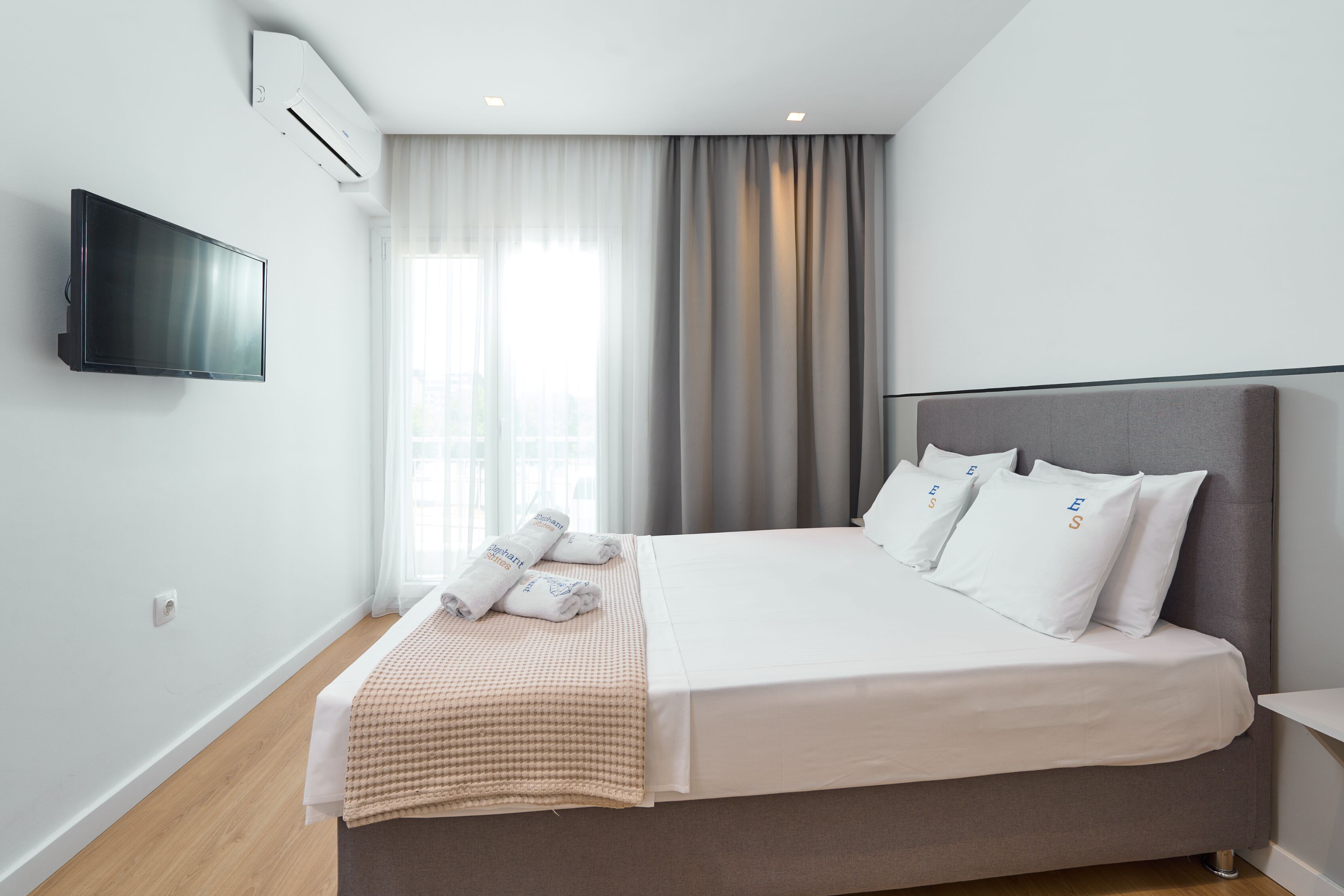A modern hotel room with a large bed, a flat-screen TV mounted on the wall, and a window with curtains.