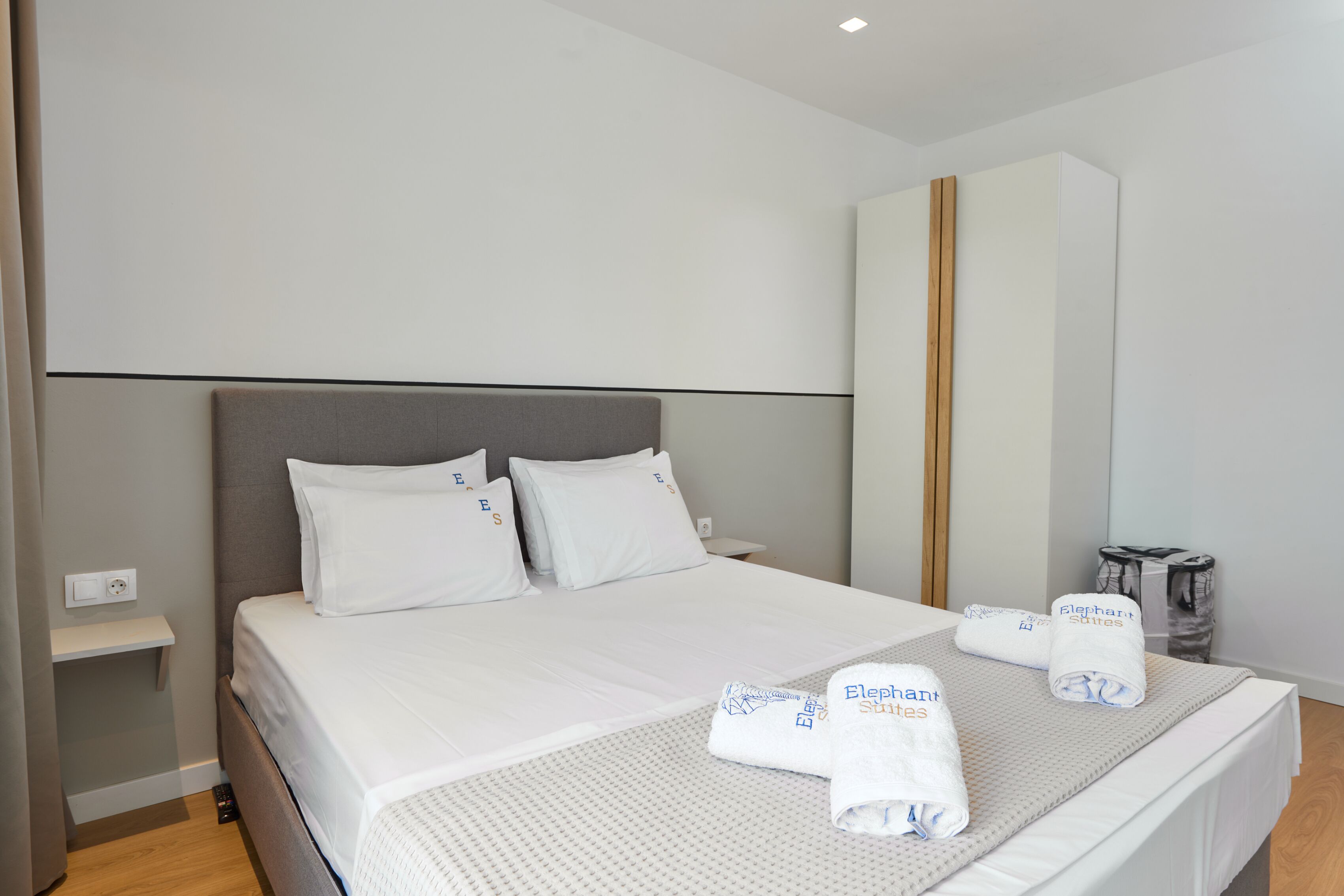 Una cama bien hecha con sábanas blancas y dos toallas con el logotipo de 'Elephant Suites', una mesita de noche y un armario grande.