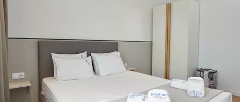 Una cama bien hecha con sábanas blancas y dos toallas con el logotipo de 'Elephant Suites', una mesita de noche y un armario grande.