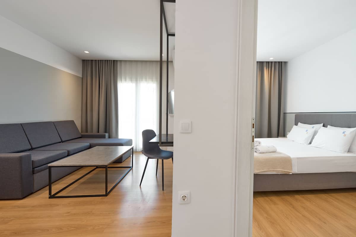Moderni hotellihuone, jossa on harmaa sohva, pieni pöytä, valkoisilla lakanoilla varustettu sänky ja puulattia.